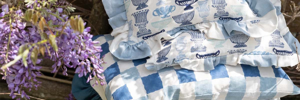 A close up image of two of molly mahons cushions with a frilly edge, made from her vases fabric design, placed on a bench underneath a blooming wisteria.