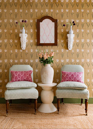 Two patterned chairs with pink cushions in a room with floral wallpaper and a small table with a vase.