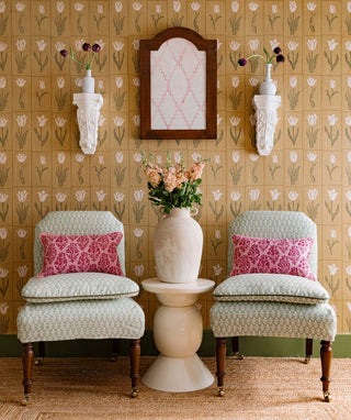 Two patterned chairs with pink cushions in a room with floral wallpaper and a small table with a vase.