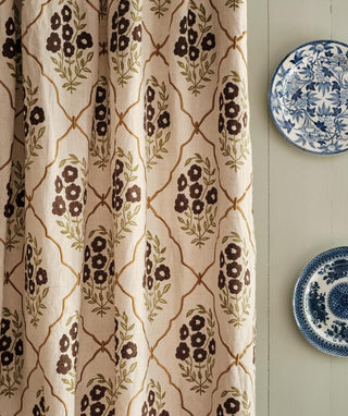 A close-up of a curtain panel with a cream background, brown floral motifs, and a tan trellis pattern, hanging next to two blue-and-white decorative plates on a light wall.