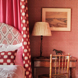 A styled bedroom corner featuring a bed with patterned headboard and pillows, a matching red and cream curtain, a wooden nightstand with a pleated lamp, a patterned wall, and a framed landscape print.