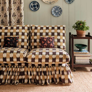 A ruffled slipcover in a brown, beige, and cream ikat pattern is draped over a sofa in a styled living room. The sofa is decorated with two small floral accent pillows, and the scene includes a wooden side table with books and a potted plant, patterned curtains, and decorative plates on the wall.