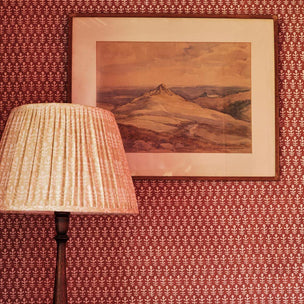 A framed landscape painting of a mountainous valley hangs on a wall with red patterned wallpaper, next to a pleated lampshade on a floor lamp.
