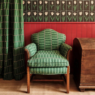 A green upholstered armchair with vertical stripe texture and cream stitching, set in a room with red wainscoting, patterned wallpaper, a dark green curtain, and a wooden chest.