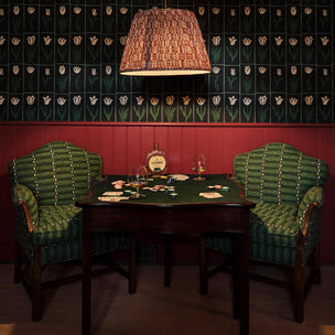 A vintage-style dark wood card table set between two green striped upholstered armchairs, under a woven pendant lamp, set against a red wainscoted wall with dark patterned wallpaper.