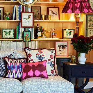 Image of a sofa in Molly Mahons Ashdown fabric in cool blue, her new collection of cushions spread across and her new ikat flirty skirt lampshade placed on a lamp standing on a side table to the right.